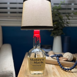 May include: A lamp made from a Maker's Mark bourbon bottle with a red wax drip and a beige lampshade with a brown trim.