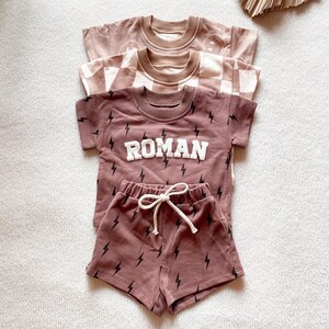 以下が含まれることがあります： 茶色の子供用Tシャツ3枚。1枚はスマイリーフェイスのデザイン、もう1枚はチェック柄、3枚目は「ROMAN」の文字と稲妻のグラフィック。稲妻模様のショートパンツも含まれています。