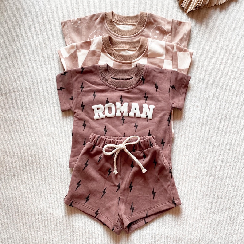 以下が含まれることがあります： 茶色の子供用Tシャツ3枚。1枚はスマイリーフェイスのデザイン、もう1枚はチェック柄、3枚目は「ROMAN」の文字と稲妻のグラフィック。稲妻模様のショートパンツも含まれています。
