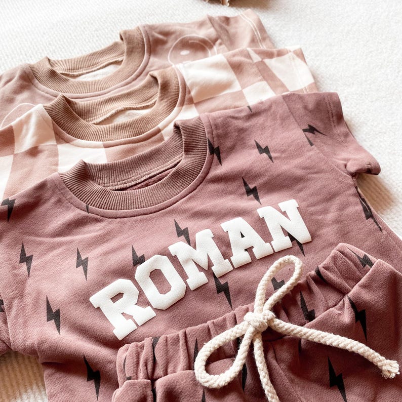 以下が含まれることがあります： 茶色の色合いの子供服の3つのマッチングセット。前面のセットは、白いブロック文字と黒い稲妻グラフィックで「ROMAN」という名前が入った、くすんだバラ色です。他の2つのセットは、チェッカーボードとスマイリーフェイスのデザインです。