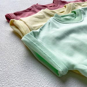 May include: Four pastel-colored short-sleeved shirts are arranged diagonally. The shirts are in shades of pink, yellow, and green. The sleeves are rolled up, revealing the inside fabric and stitching. The shirts are made of a soft material.