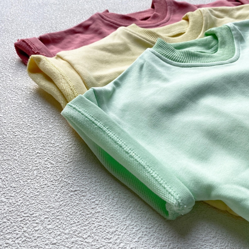May include: Four pastel-colored short-sleeved shirts are arranged diagonally. The shirts are in shades of pink, yellow, and green. The sleeves are rolled up, revealing the inside fabric and stitching. The shirts are made of a soft material.