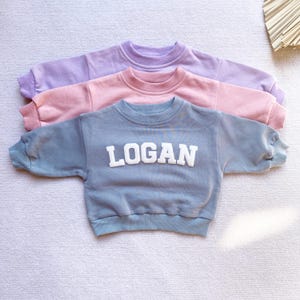 May include: Three sweatshirts stacked on top of each other. The top sweatshirt is lavender, the middle sweatshirt is pink, and the bottom sweatshirt is light blue. The bottom sweatshirt has the name "LOGAN" in white letters on the front.