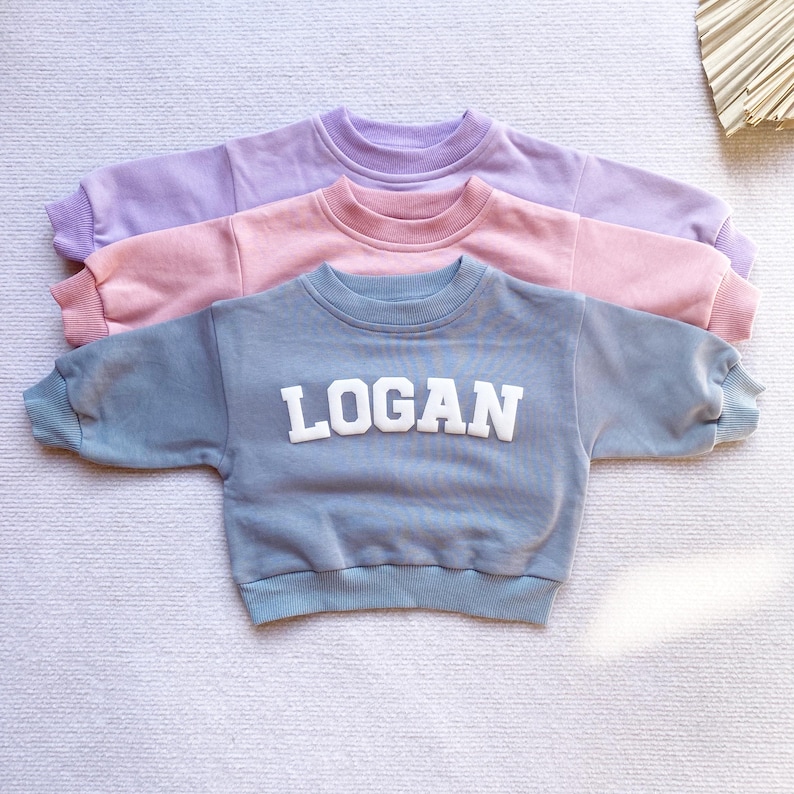 May include: Three sweatshirts stacked on top of each other. The top sweatshirt is lavender, the middle sweatshirt is pink, and the bottom sweatshirt is light blue. The bottom sweatshirt has the name "LOGAN" in white letters on the front.