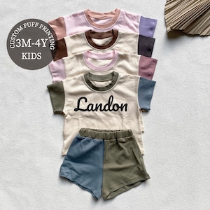 May include: A stack of children's t-shirts and shorts sets in various colors. The t-shirts have contrasting sleeves and necklines, with the name "Landon" printed on one. The shorts are two-toned. The image includes text that says "Custom Puff Printing" and "3M-4Y KIDS".
