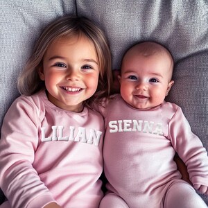 May include: Two young girls wearing pink long-sleeved shirts with white embroidered names. The older girl's shirt says "Lilian" and the younger girl's shirt says "Sienna".