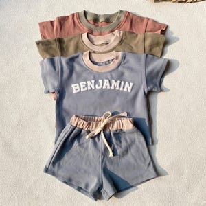 Puede incluir: Un conjunto de dos piezas azul y rosa para un niño. La parte superior es una camiseta de manga corta con cuello de doble capa y la palabra "BENJAMIN" en letras blancas. La parte inferior es un pantalón corto con una cintura rosa.