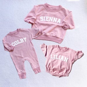May include: Three pink baby outfits with white lettering. The first outfit is a pink sweatshirt with the name "SIENNA" in white letters. The second outfit is a pink onesie with the name "COLBY" in white letters. The third outfit is a pink sweatshirt with the name "LILIAN" in white letters.