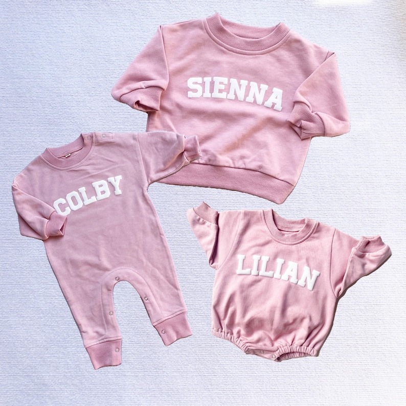 May include: Three pink baby outfits with white lettering. The first outfit is a pink sweatshirt with the name "SIENNA" in white letters. The second outfit is a pink onesie with the name "COLBY" in white letters. The third outfit is a pink sweatshirt with the name "LILIAN" in white letters.