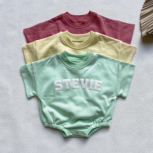 May include: Three baby rompers in pastel colors: dusty rose, pale yellow, and mint green. The mint green romper features the name "STEVIE" in white block letters. Each romper has short sleeves and a round neckline.