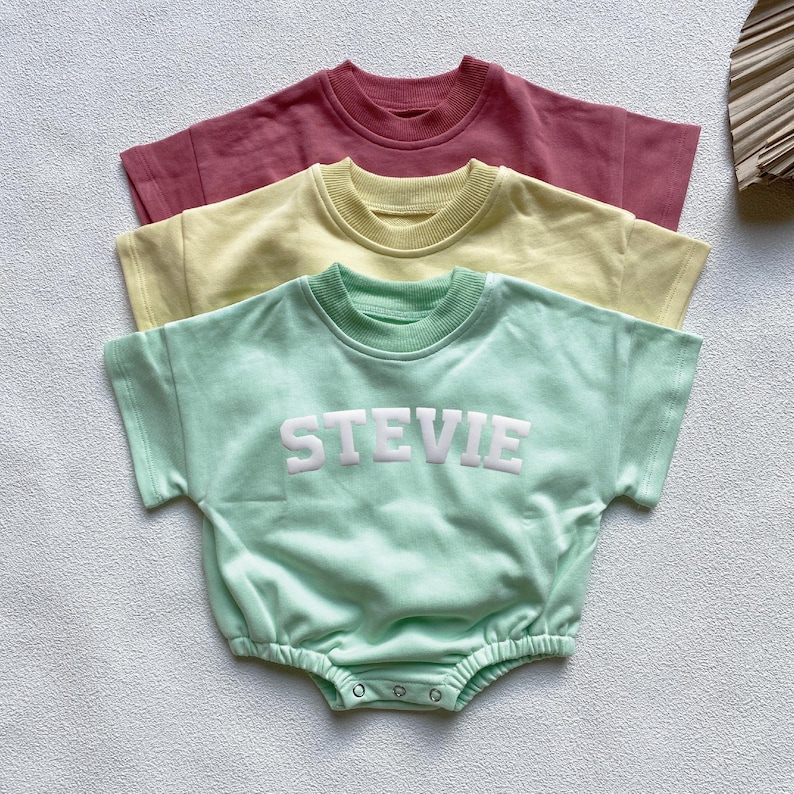 May include: Three baby rompers in pastel colors: dusty rose, pale yellow, and mint green. The mint green romper features the name "STEVIE" in white block letters. Each romper has short sleeves and a round neckline.