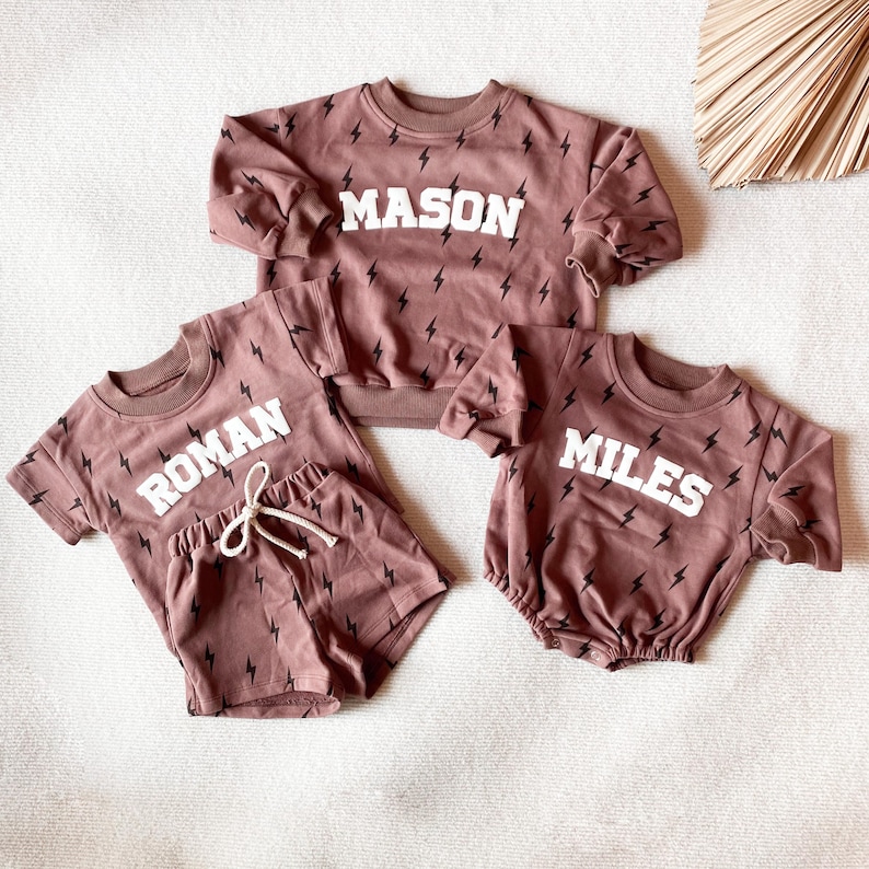 以下が含まれることがあります： モーヴ色の子供服の3つのマッチングセット。それぞれに黒い稲妻模様が施されています。「MASON」の白いブロック文字が入ったスウェットシャツのセットが1つ。もう1つのセットには、「ROMAN」のシャツとショーツがあります。3つ目のセットには「MILES」のロンパースがあります。