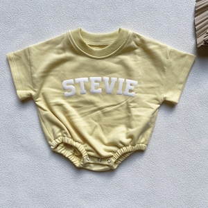 May include: A light yellow baby romper with short sleeves and the name "STEVIE" in white block letters across the chest. The romper has a round neckline and snaps at the bottom. The fabric appears soft and comfortable.