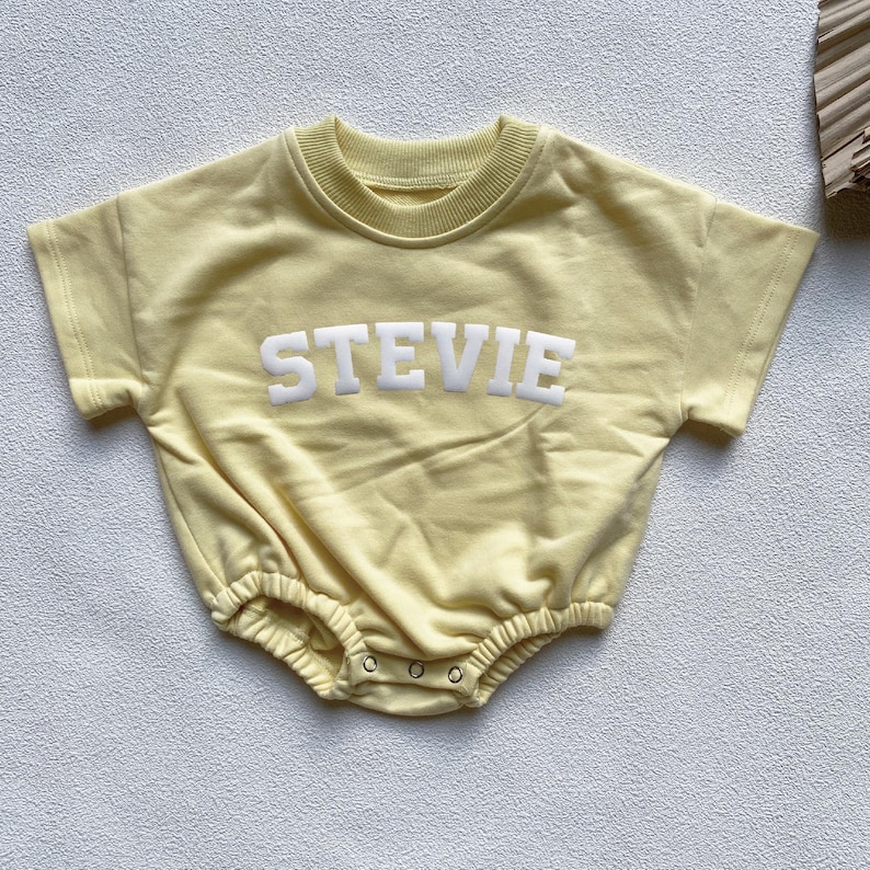 May include: A light yellow baby romper with short sleeves and the name "STEVIE" in white block letters across the chest. The romper has a round neckline and snaps at the bottom. The fabric appears soft and comfortable.