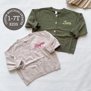 May include: Two children's sweaters. One is light pink with "Reyna" embroidered in red, the other is olive green with "Liam" embroidered in yellow. Both have button closures and ribbed cuffs. The image includes a circular graphic that says "Custom Matching Sweater 1-7T Kids."