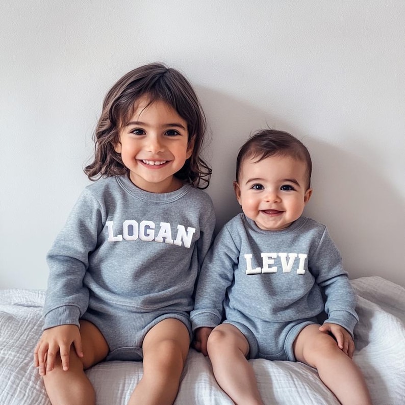 May include: Two children wearing grey sweatshirts with their names, Logan and Levi, embroidered in white on the front. The children are sitting on a white textured surface and smiling at the camera.