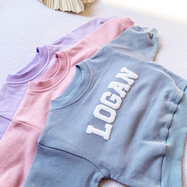 May include: Three sweatshirts in different colors: lavender, pink, and light blue. The light blue sweatshirt has the name "LOGAN" printed in white on the front.