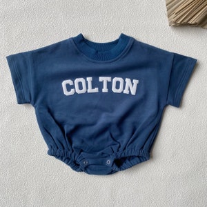 May include: Navy blue baby romper with the name "COLTON" in white block letters across the chest. The romper has short sleeves, a round neckline, and a gathered waist. The bottom has snap closures.