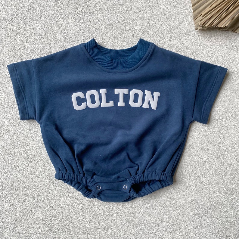 May include: Navy blue baby romper with the name "COLTON" in white block letters across the chest. The romper has short sleeves, a round neckline, and a gathered waist. The bottom has snap closures.