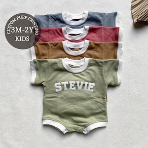 Può includere: Quattro tutine per bambini in tonalità blu, rosso, marrone e verde, ciascuna con colletto e bordi delle maniche bianchi. La tutina verde presenta il nome "STEVIE" a lettere bianche. L'immagine include il testo "CUSTOM PUFF PRINTING 3M-2Y KIDS."
