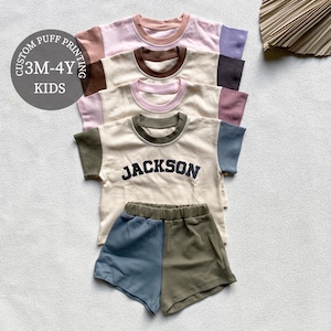 May include: A stack of children's t-shirts and shorts sets in various colors, including pink, cream, and olive green. The shirts have contrasting sleeves and necklines, with the word "JACKSON" printed on one. The sets are for ages 3M-4Y.