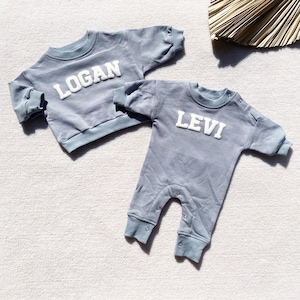 May include: Two baby outfits in a light blue color. One is a long-sleeved shirt with the name "LOGAN" in white letters. The other is a one-piece jumpsuit with the name "LEVI" in white letters.