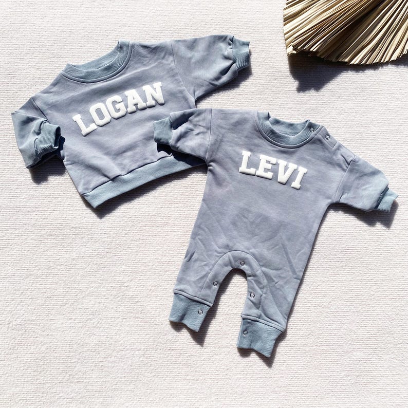 May include: Two baby outfits in a light blue color. One is a long-sleeved shirt with the name "LOGAN" in white letters. The other is a one-piece jumpsuit with the name "LEVI" in white letters.