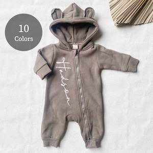 Pode incluir: Um macacão de bebê marrom com capuz, orelhas de urso e fecho de zíper. O macacão tem o nome "Hudson" bordado em branco na frente.