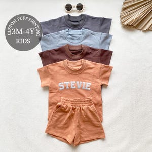 May include: Four short-sleeved children's shirts in grey, light blue, brown, and peach. The peach shirt has the name "STEVIE" in white letters. A matching peach shorts set is below the peach shirt. Sunglasses are at the top.