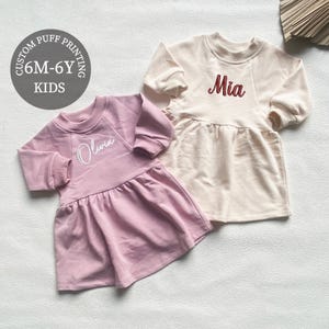 Könnte beinhalten: Zwei Kinderkleider, eines hellrosa und eines cremefarben, mit langen Ärmeln und einem gerafften Rock. Das rosa Kleid hat "Olivia" in Weiß gestickt, und das cremefarbene Kleid hat "Mia" in Rot gestickt. Die Kleider sind für Kinder im Alter von 6 Monaten bis 6 Jahren.