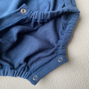 May include: Close-up of a navy blue baby romper made of soft, textured fabric. The romper features elasticated waist and leg openings, and snap closures. The garment is designed for comfort and ease of wear.