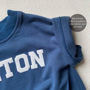 May include: Close-up of a navy blue sweatshirt with white block lettering. The sweatshirt has a crew neck and a thick material, suitable for spring and autumn wear. The text on the sweatshirt is partially visible.