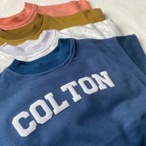 May include: A stack of crew neck sweatshirts in various colors: navy blue with white text, light grey, olive green, and peach. The navy sweatshirt features the name "COLTON" in bold, white, block letters.