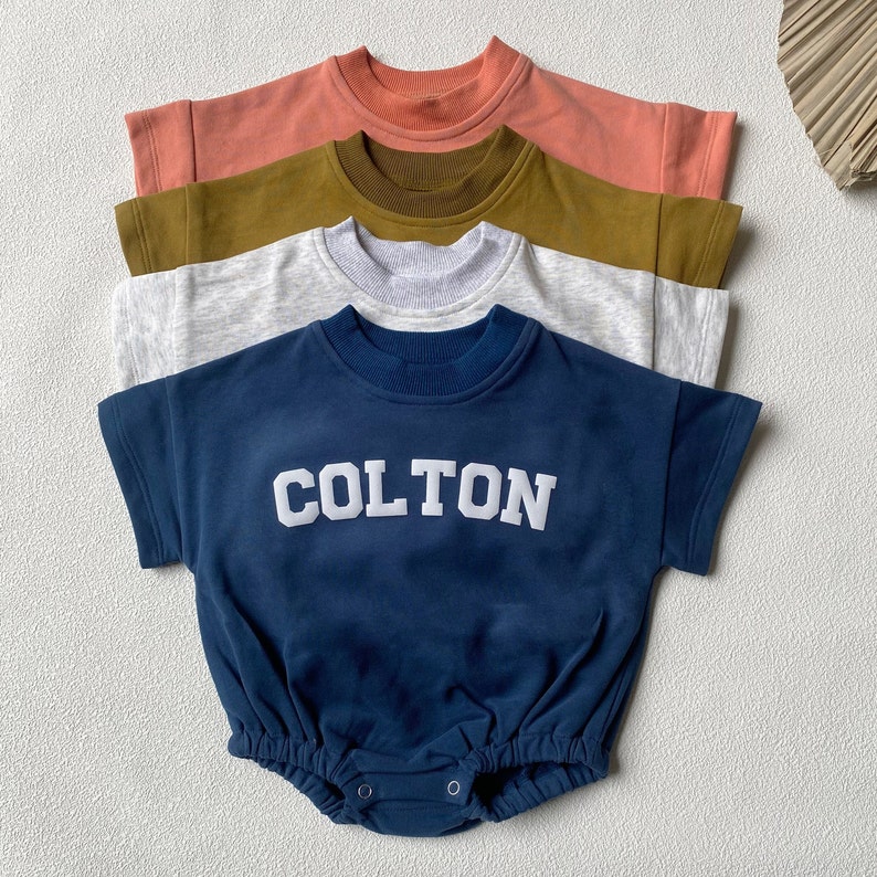 May include: Four baby rompers in a row, each with short sleeves and a round neckline. The colors are coral, olive green, light gray, and navy blue. The navy blue romper has the name "COLTON" in white block letters across the chest.