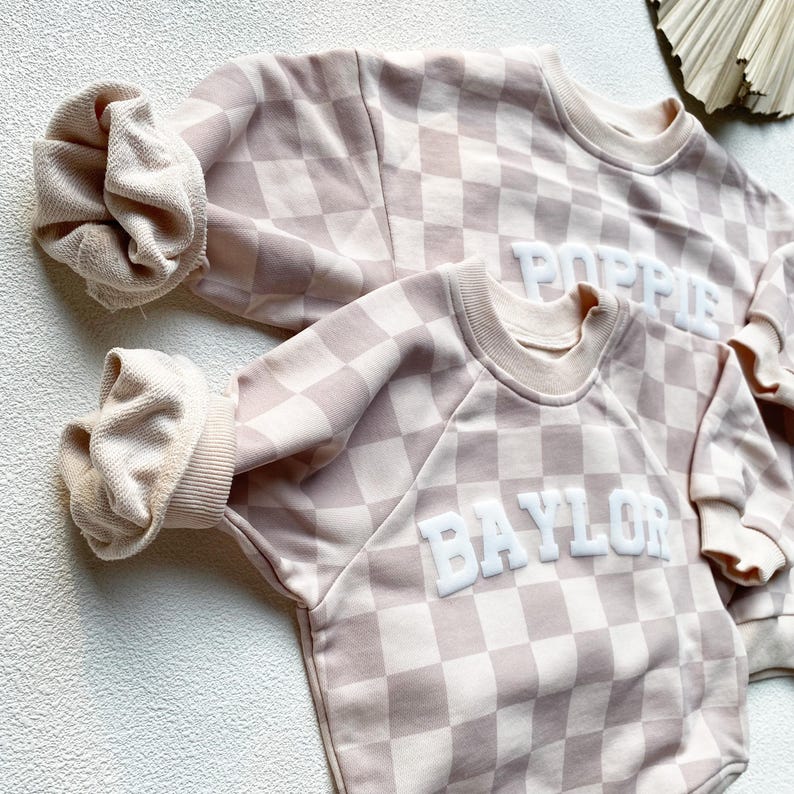 May include: Two matching light pink and white checkered sweatshirts. One sweatshirt has the name "POPPY" in white letters, and the other has "BAYLOR" in white letters. The sweatshirts have ribbed cuffs and a crew neck.