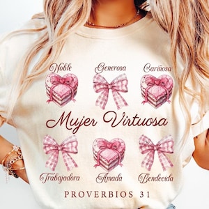 May include: A white t-shirt with a pink and brown design featuring a heart-shaped cake with the words "Mujer Virtuosa" and "Proverbios 31" in brown text. The design also includes the words "Noble", "Generosa", "Cari&ntilde;osa", "Trabajadora", "Amada", and "Bendecida" in pink text.