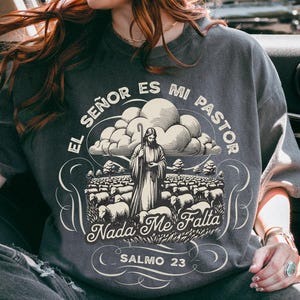 May include: Dark grey t-shirt featuring a graphic of a shepherd and sheep, with the text "EL SEÑOR ES MI PASTOR, NADA ME FALTA, SALMO 23". The shirt is worn by a person wearing sunglasses and a brown hat.