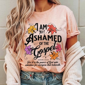 May include: A pink t-shirt with a floral design and the text "I AM NOT ASHAMED OF THE GOSPEL" with the scripture reference "ROMANS 1:16 KJV" and the text "For it is the power of God unto salvation for everyone that believeth."