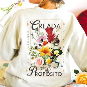 May include: A cream-colored long-sleeve sweatshirt featuring a back graphic of a colorful floral bouquet. The text reads CREADA Con Un PROPOSITO and ROMANOS 8:28, framed by four small star icons. The design is centered on the back of the garment.