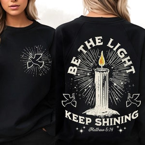 Puede incluir: Sudadera negra con un gráfico de paloma blanca en la parte delantera y un gráfico de vela en la parte trasera. El gráfico de la parte trasera dice "Be the Light Keep Shining Matthew 5:14".