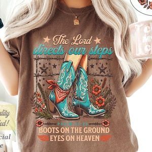 May include: Brown t-shirt with a graphic featuring turquoise cowboy boots, barbed wire, and floral accents. Text includes "The Lord directs our steps" and "Boots on the ground, eyes on heaven." A small circle with text "Psalm 37:23" is also visible.