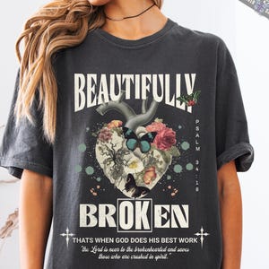 May include: A dark gray t-shirt with a graphic design of a heart with flowers and butterflies. The text "Beautifully Broken" is printed on the shirt, along with the text "Psalm 34:18" and a quote about God's work.