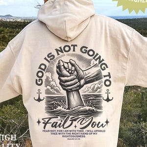 May include: A cream-colored hoodie with a graphic print on the back that says "God is not going to fail you." The graphic features a hand reaching out of the water and being grasped by another hand. The text "Fear not, for I am with thee, I will uphold thee with the right hand of my righteousness. Isaiah 41:10" is printed below the graphic.