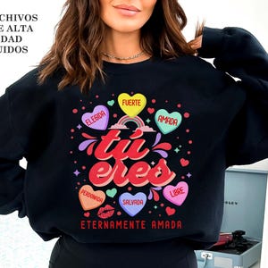 May include: Black sweatshirt with a graphic design of hearts and the text "tu eres" in Spanish, which translates to "you are". The hearts have different colors and each has a word written on it, such as "elegida" (chosen), "fuerte" (strong), and "amada" (loved). The text "eternamente amada" (eternally loved) is written below the graphic.