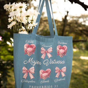 May include: A light blue tote bag with a pink and white design. The bag features the text "Mujer Virtuosa" and "Proverbios 31" along with illustrations of hearts and bows. The text "Noble, Generosa, Cari&ntilde;osa, Trabajadora, Amada, Bendicida" is also included.