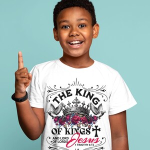May include: White t-shirt with a black graphic design that says "The King of Kings and Lord of Lords Jesus 1 Timothy 6:15". The graphic features a crown, doves, and roses.