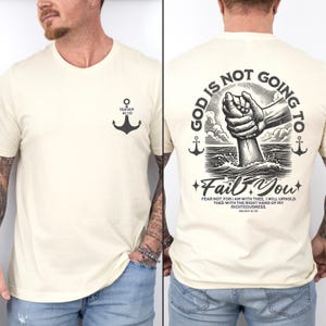 Op de afbeelding: Een cr&egrave;mekleurig T-shirt met een zwarte ankergrafiek op de linkerborst en een zwarte grafiek op de rug die zegt "God zal je niet in de steek laten" met een zwart-witte illustratie van een hand die naar beneden reikt om iemand in het water te helpen. De tekst bevat ook de Bijbeltekst Jesaja 41:10.