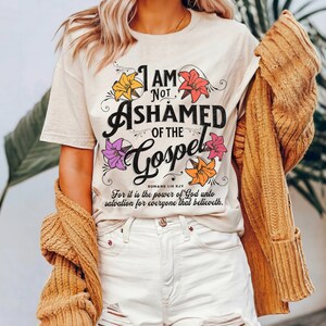 May include: A white t-shirt with a floral design and the text "I am not ashamed of the Gospel" with the scripture reference "Romans 1:16 KJV" and the text "For it is the power of God unto salvation for everyone that believeth."