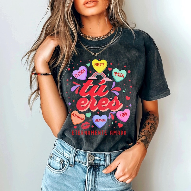 May include: A black t-shirt with a graphic design of hearts and the text "tu eres" in a colorful font. The hearts have Spanish words on them, including "elegida", "fuerte", "amada", "perdonada", "salvada", and "libre". The text at the bottom of the design reads "eternamente amada".