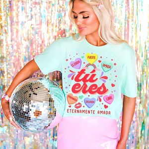 May include: A light blue t-shirt with a graphic design featuring the Spanish words "tu eres" in a colorful font. The graphic also includes hearts with Spanish words like "elegida", "fuerte", "amada", "perdonada", "salvada", and "libre". The shirt also has the text "Eternamente Amada" at the bottom.